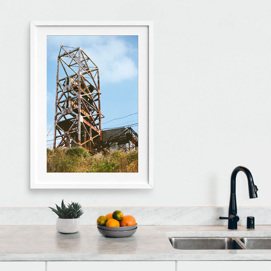 Rusted steel lattice of a mine headframe at Ikeshima, with power lines crossing toward a corrugated shed and dry golden grass on the slope below.