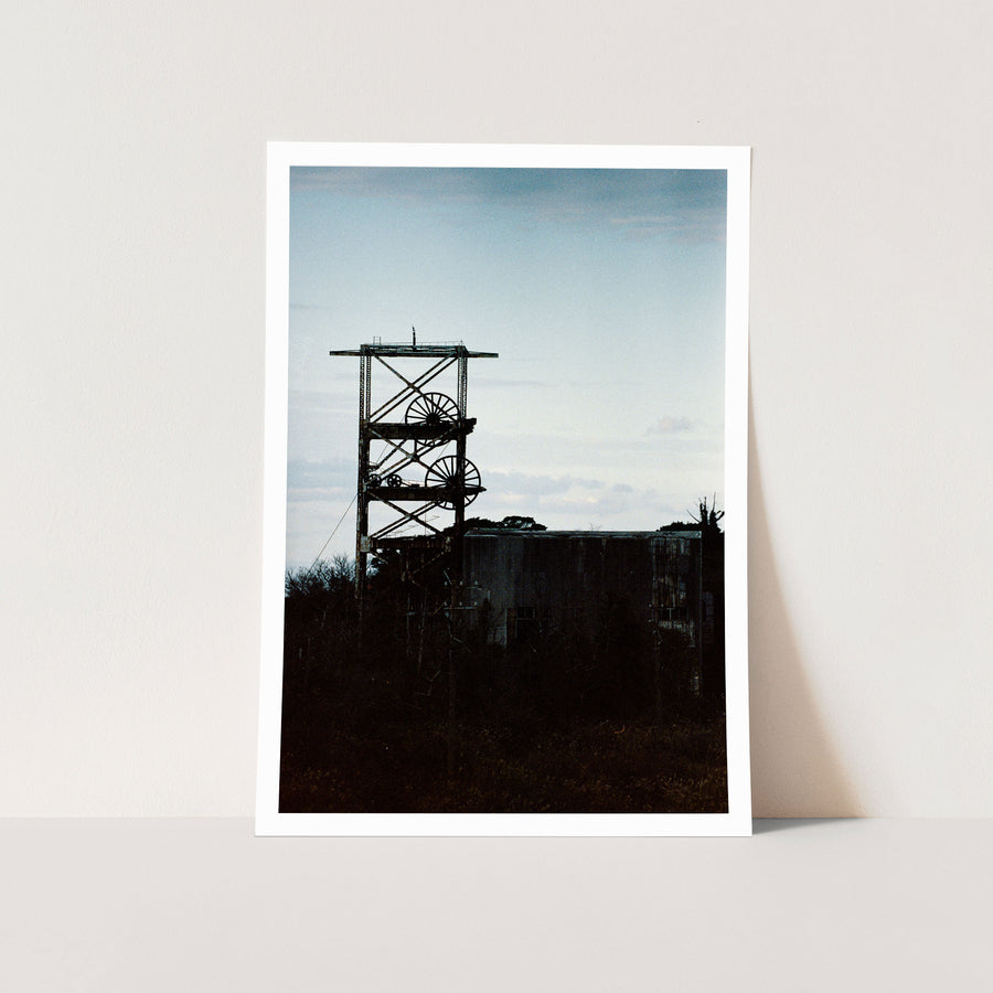 The twin spoked winding wheels of the steel lattice headframe at Ikeshima Coal Mine, silhouetted against a warm sunset sky, with dense foliage surrounding the lower framework and a corrugated metal building visible to the right.