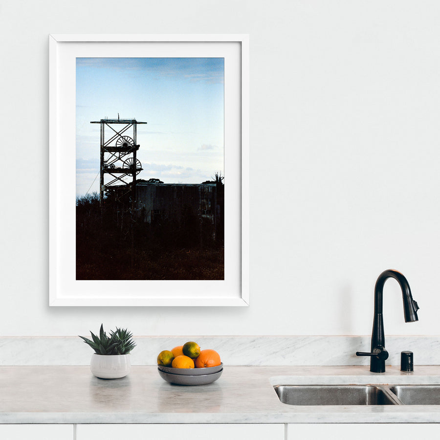 The twin spoked winding wheels of the steel lattice headframe at Ikeshima Coal Mine, silhouetted against a warm sunset sky, with dense foliage surrounding the lower framework and a corrugated metal building visible to the right.