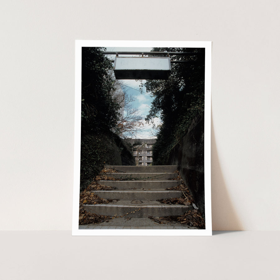 Concrete steps rise between ivy-covered and bare retaining walls at Ikeshima, a worn white sign frame hanging above the lower treads and power lines crossing a blue sky overhead.