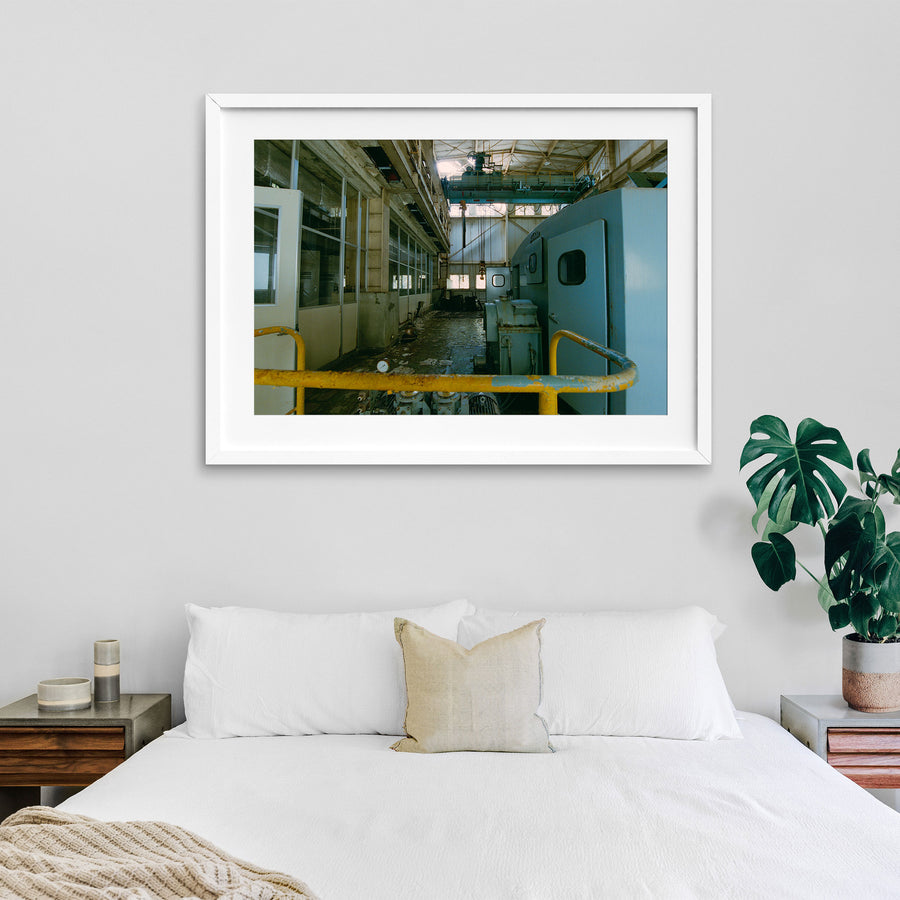 Interior of the winding house at Ikeshima Coal Mine, showing a light-blue winding engine with panelled doors, a teal overhead gantry crane, glass-paned windows, and a pressure gauge on the concrete floor.
