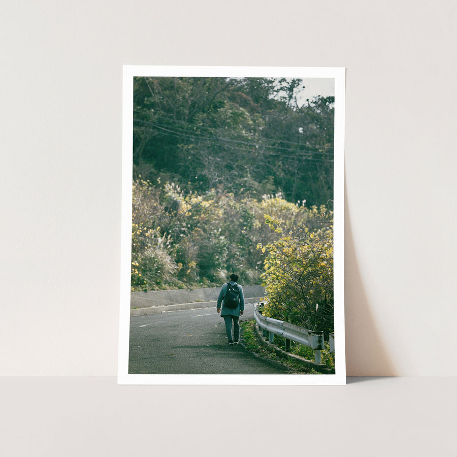 A person in a light blue coat walks away up a curving road on Ikeshima, his long shadow stretched across the asphalt and yellow-green scrub crowding the rusted guardrail to the right.