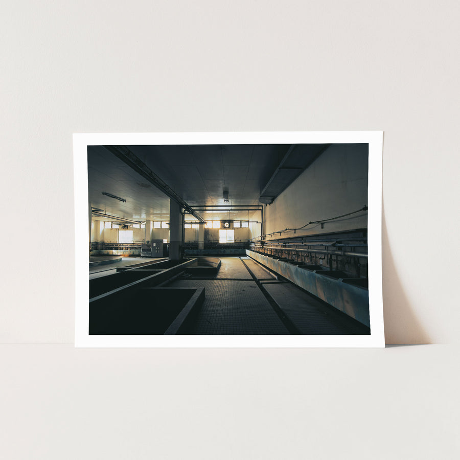 Interior of the miners' bathhouse at Ikeshima Coal Mine, with a clock above far windows, concrete washing troughs across the tiled floor, and a rusted spigot trough along the right wall.
