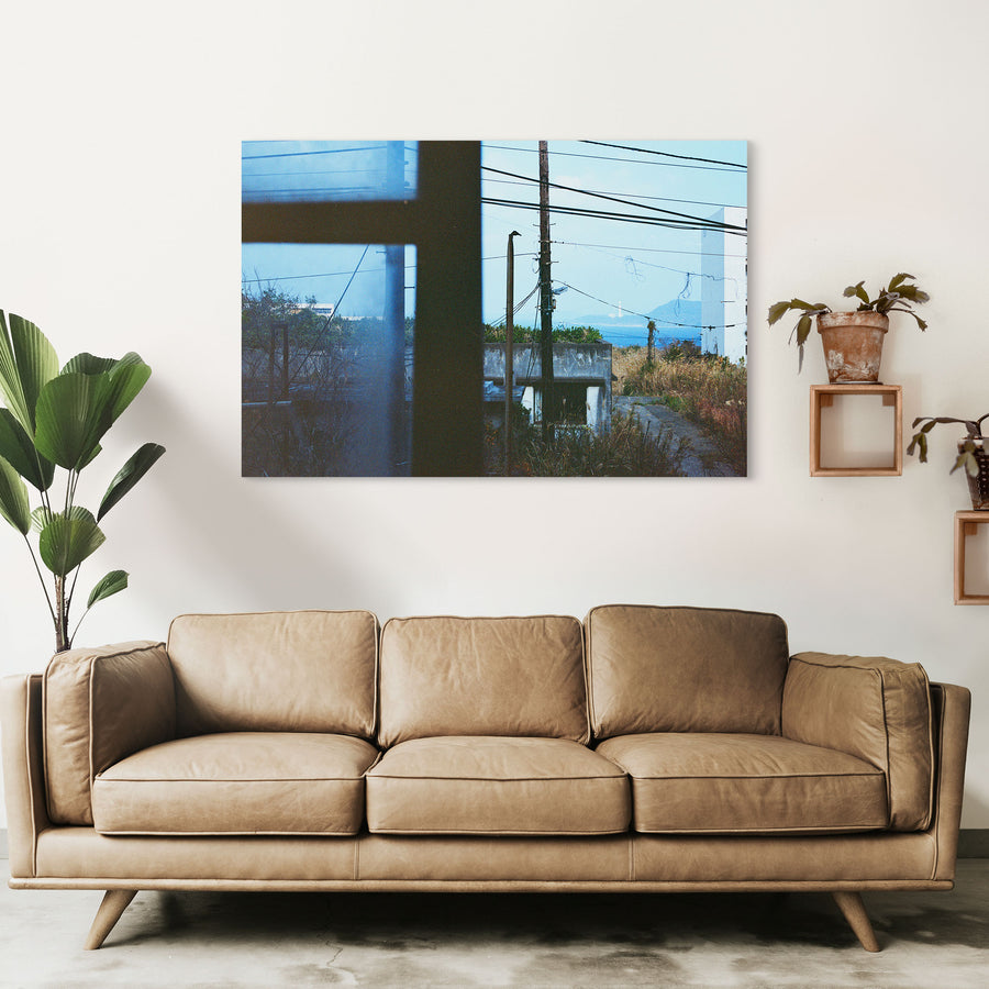 View through a dark window frame at Ikeshima showing two utility poles with tangled wires against pale sky, dry grass, a concrete structure, and a white lighthouse on a distant landmass across open water.