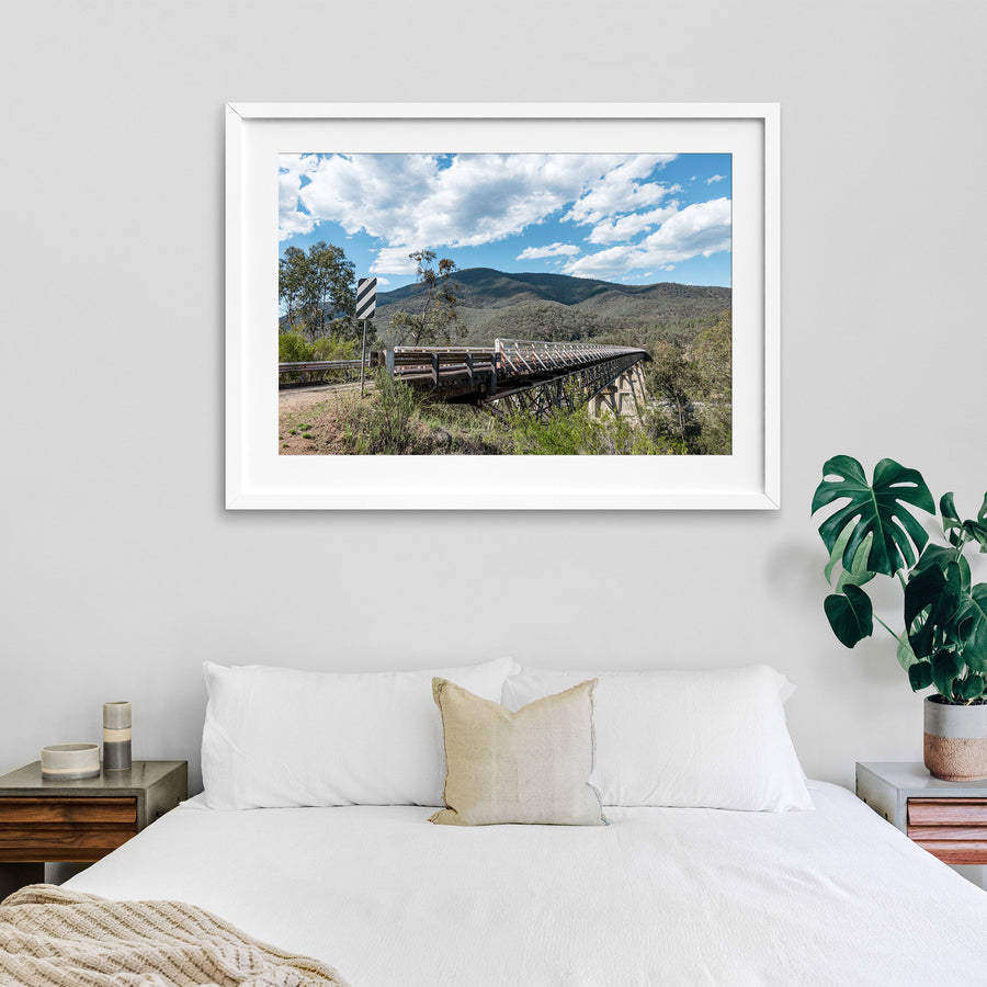 View along the road approach to McKillops Bridge in Deddick Valley, with a black and white chevron sign at left and the single-lane timber-decked steel truss bridge spanning the Snowy River, forested hills rising beyond under an overcast sky.