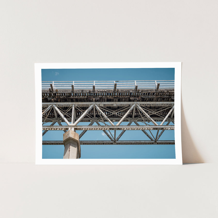 View from below McKillops Bridge in Deddick Valley, looking up at the electric-arc-welded steel Warren trusses and weathered timber decking against a clear sky, with a tall concrete pier at the left.