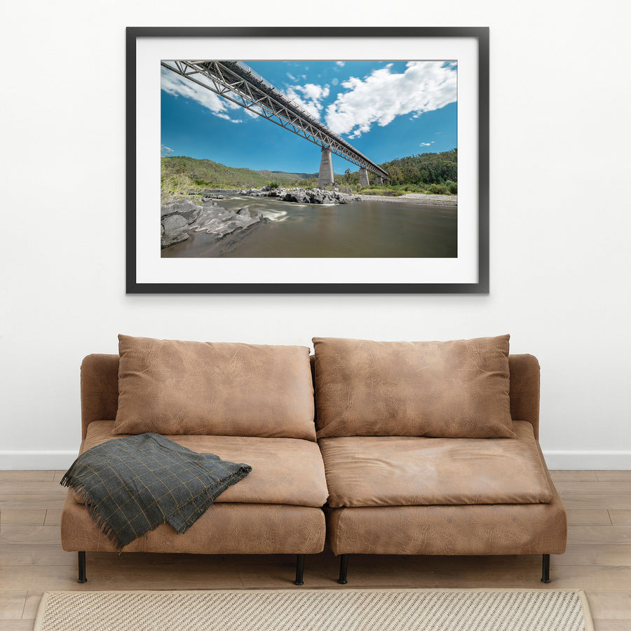 View from the rocky riverbed looking up at the steel Warren trusses and concrete A-frame pier of McKillops Bridge in the Deddick Valley, with the wide brown Snowy River running over exposed rock below and wooded hills behind.