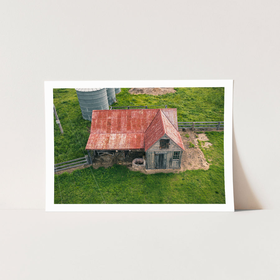 Aerial view of a timber cottage and shed under a faded red corrugated iron roof at Jembaicumbene, with a pig beside a stone trough near the doorway and two metal silos on the surrounding paddock.