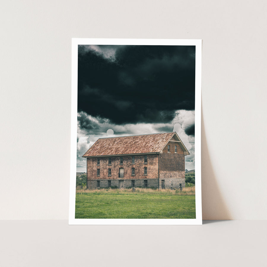 The four-storey brick and granite flour mill at Jembaicumbene, its multi-paned windows and central iron balcony visible against a heavy grey sky, photographed in open grassland near Braidwood.