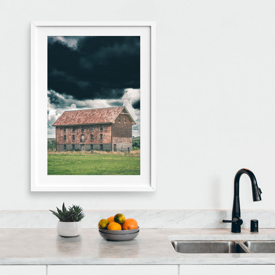 The four-storey brick and granite flour mill at Jembaicumbene, its multi-paned windows and central iron balcony visible against a heavy grey sky, photographed in open grassland near Braidwood.