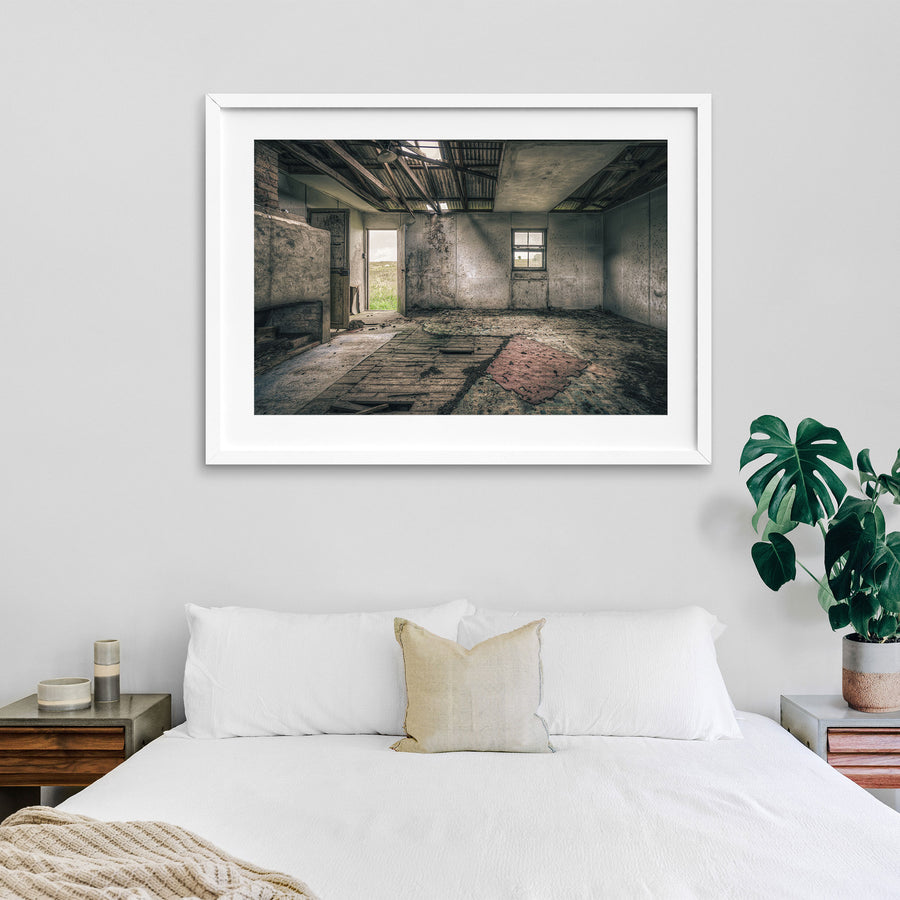 Interior of a derelict room at Jembaicumbene with a partly collapsed corrugated metal roof open to the sky, exposed timber rafters, rendered walls, a four-pane sash window, an open timber door to a paddock, and a torn red rug on broken floorboards.