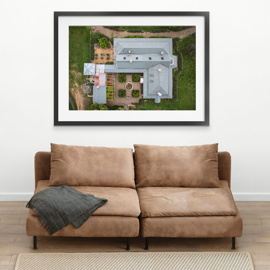Aerial view of the formal gravel courtyard at Mill Pond Farm, Jembaicumbene, with a circular stone fountain at centre, clipped hedges, and grey corrugated metal roofs surrounding on three sides.