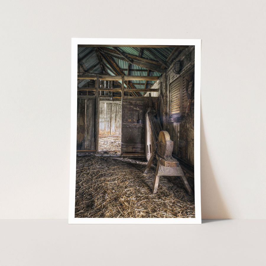 A large stone wheel on a timber frame inside a weathered barn at Jembaicumbene, with a plank door, louvred window, straw-covered dirt floor, and corrugated iron roof on rough timber beams.