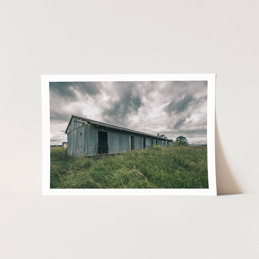A weathered corrugated iron building with empty window openings and an open doorway stands in tall grass at Jembaicumbene, New South Wales.