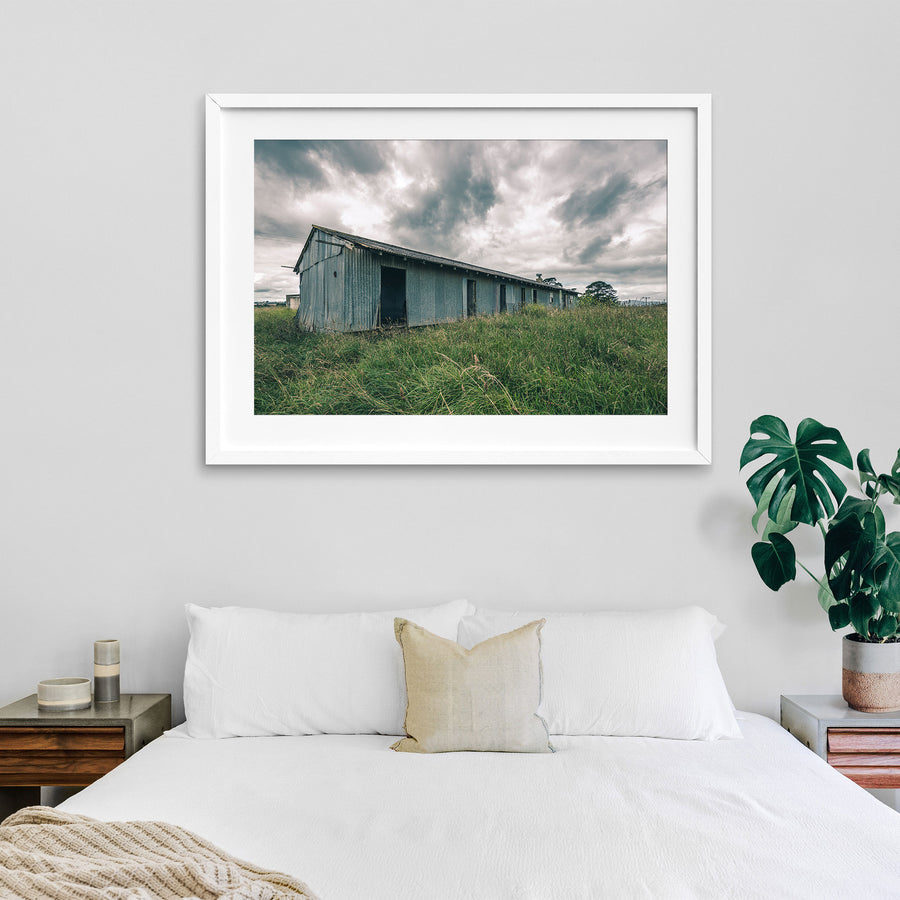 A weathered corrugated iron building with empty window openings and an open doorway stands in tall grass at Jembaicumbene, New South Wales.