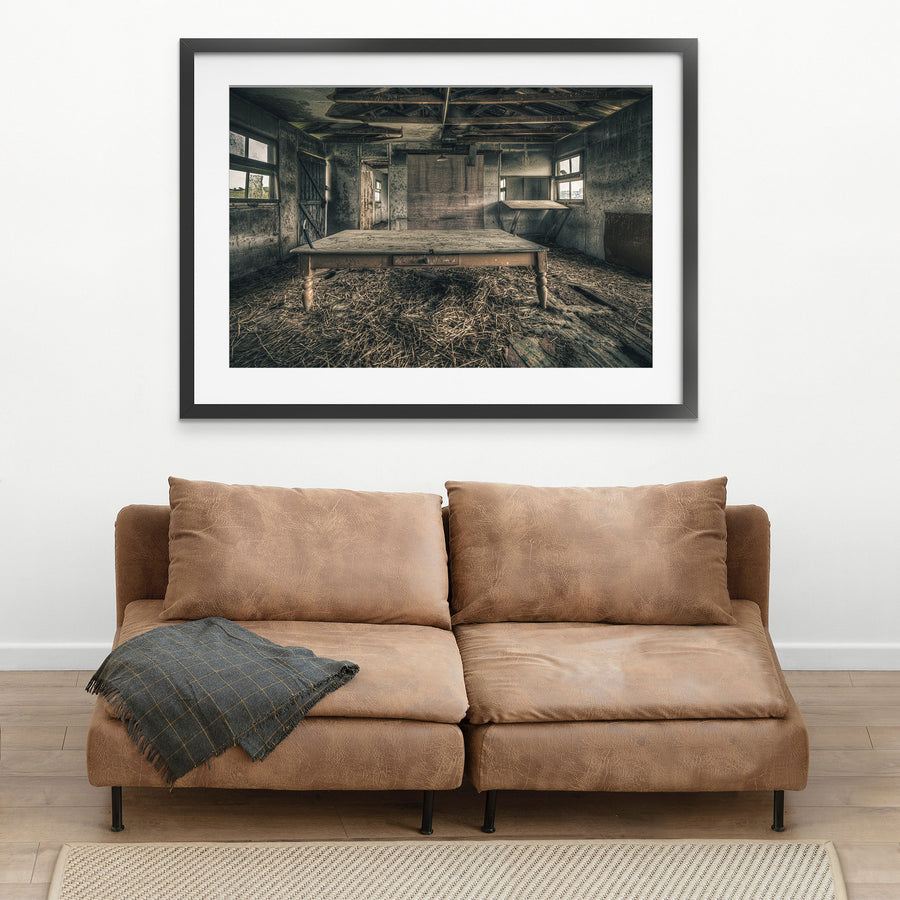 A long timber table with turned legs stands on a straw-strewn floor inside a derelict outbuilding at Mill Pond Farm, Jembaicumbene, with exposed rafters overhead and light entering from windows on either side.