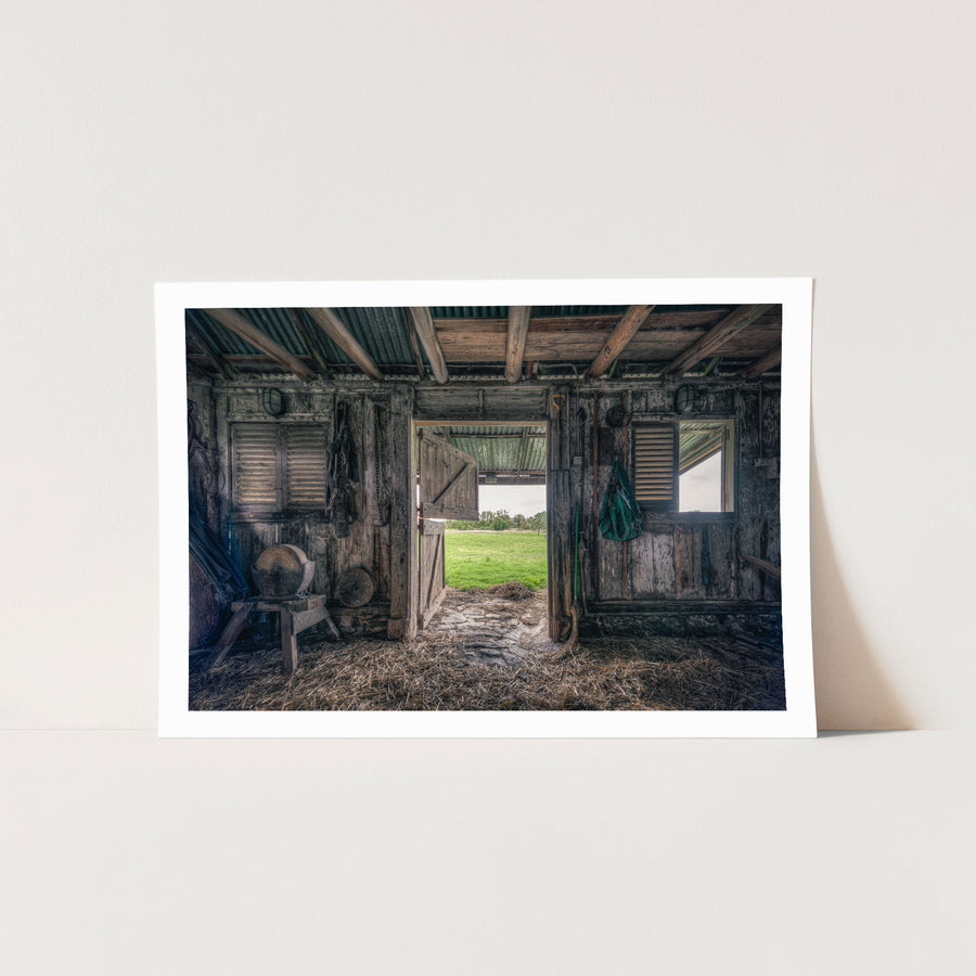 Interior of the 1840s timber wagon barn at Jembaicumbene, looking through an open doorway to a green paddock and treeline, with a chain and hay net hanging from weathered posts and straw on the dirt floor.