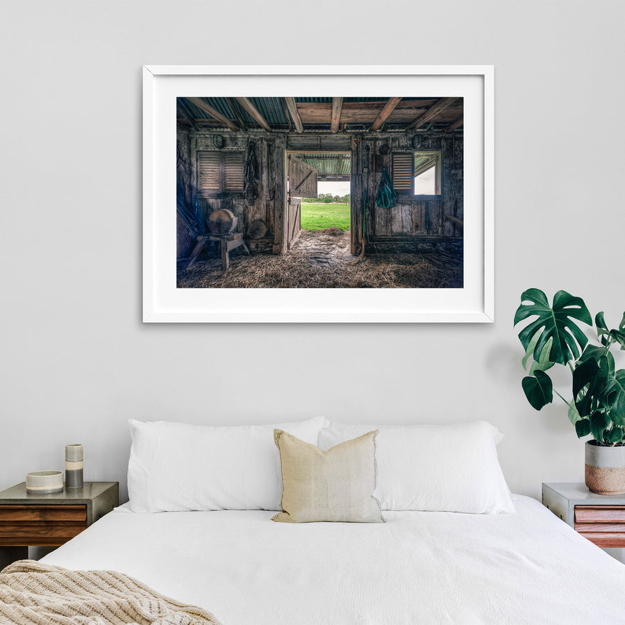 Interior of the 1840s timber wagon barn at Jembaicumbene, looking through an open doorway to a green paddock and treeline, with a chain and hay net hanging from weathered posts and straw on the dirt floor.