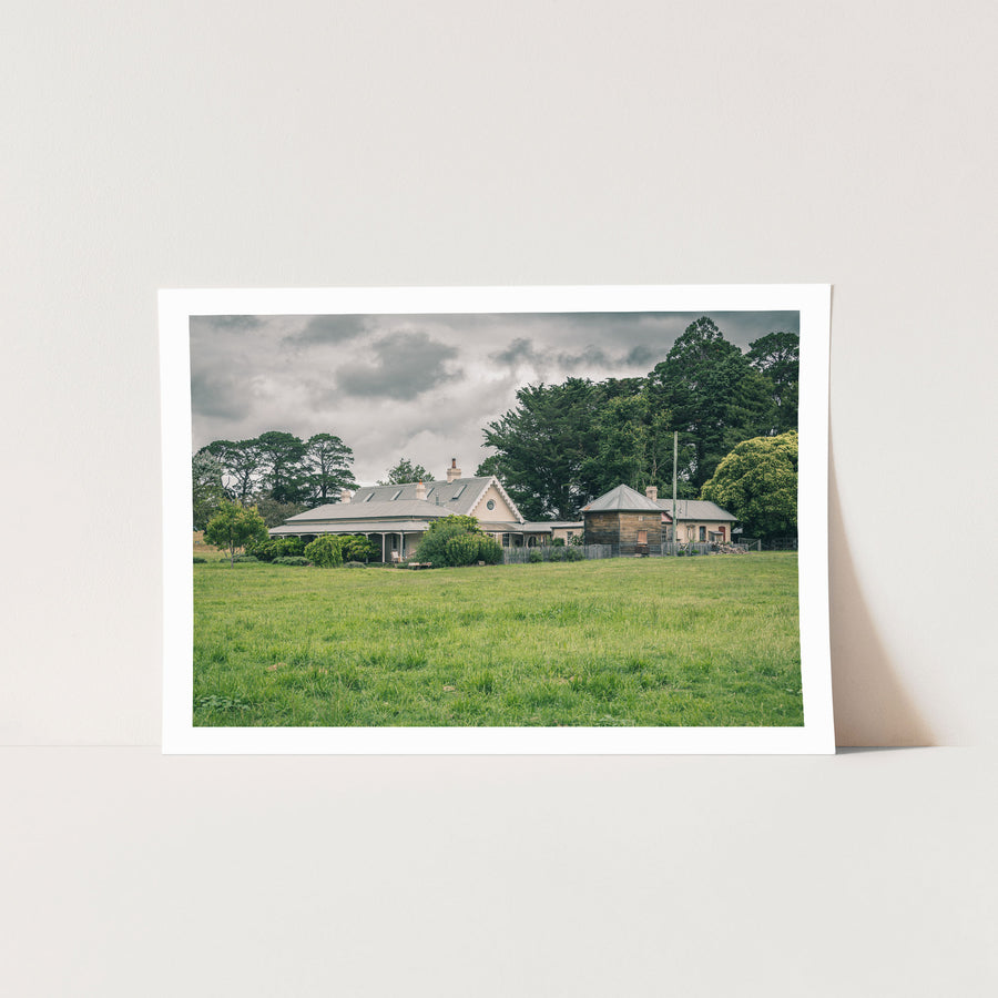 A weatherboard homestead with Gothic Revival bargeboards and a corrugated iron roof, a timber outbuilding to the right, and a broad green paddock in the foreground at Jembaicumbene, NSW.