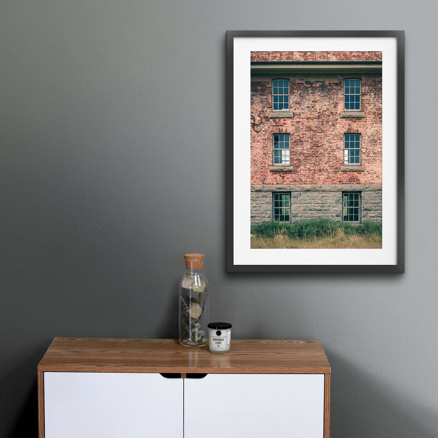 Six multi-pane sash windows set in three pairs across a brick and stone facade at Jembaicumbene, with corrugated metal roofing above and tall grass and purple flowering plants at the base.
