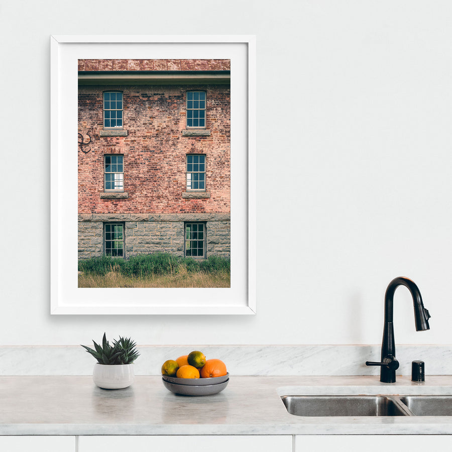 Six multi-pane sash windows set in three pairs across a brick and stone facade at Jembaicumbene, with corrugated metal roofing above and tall grass and purple flowering plants at the base.