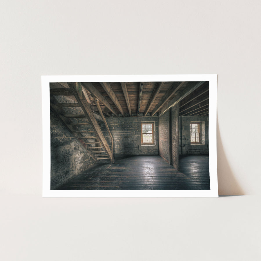 A wooden staircase rises along the stone wall of a timber-floored room at Jembaicumbene, with two double-hung windows in the rear wall looking out to open ground.