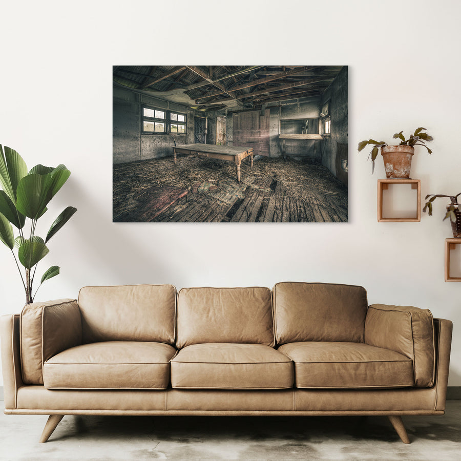 A long timber table with turned legs on a straw-strewn floor of broken boards, with multi-pane windows and an open plank door visible in a derelict interior at Jembaicumbene, New South Wales.