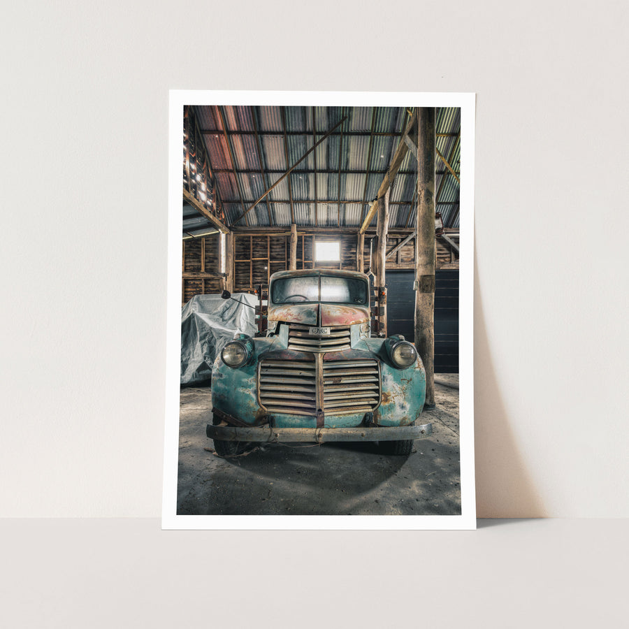 An old truck with a faded blue and rust body parked front-on inside the timber wagon barn at Jembaicumbene, its chrome grille and round headlights lit by a small window above.