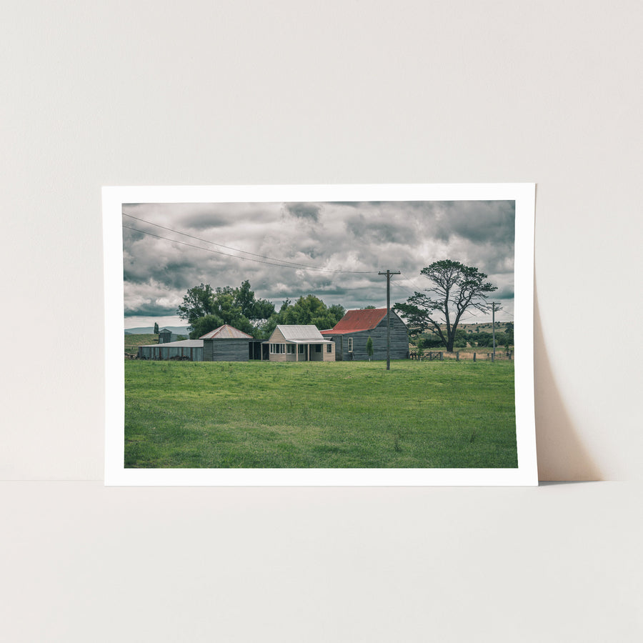 Farm buildings at Jembaicumbene, a red iron roofed barn and timber cottage across a green paddock.