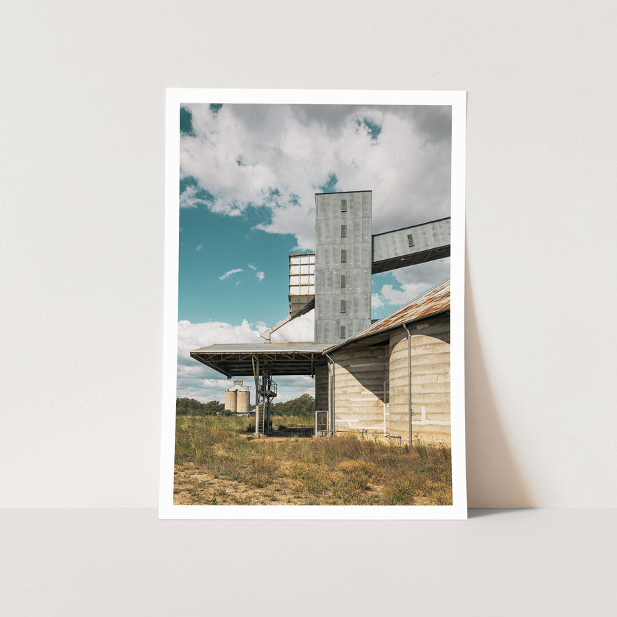 A tall corrugated metal grain elevator tower connected by a raised conveyor gallery and steel stairway to a concrete grain shed at Mount Russell, with smaller silos visible across a grassed paddock under a partly clouded sky.