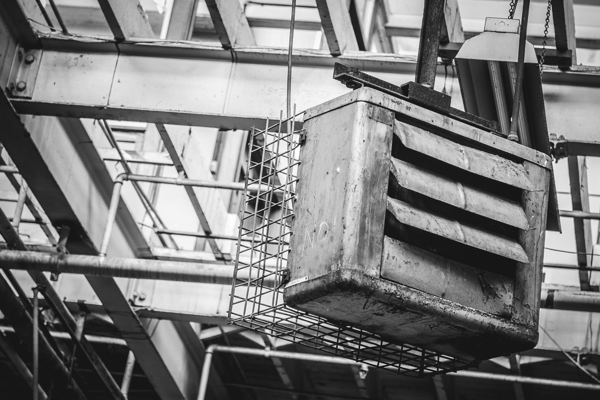 Rust and peeling paint cover a suspended industrial unit, its louvred vents clogged with grime and a wire mesh cage bolted to its underside at O-I Glass. Rough lettering is scrawled across the casing.