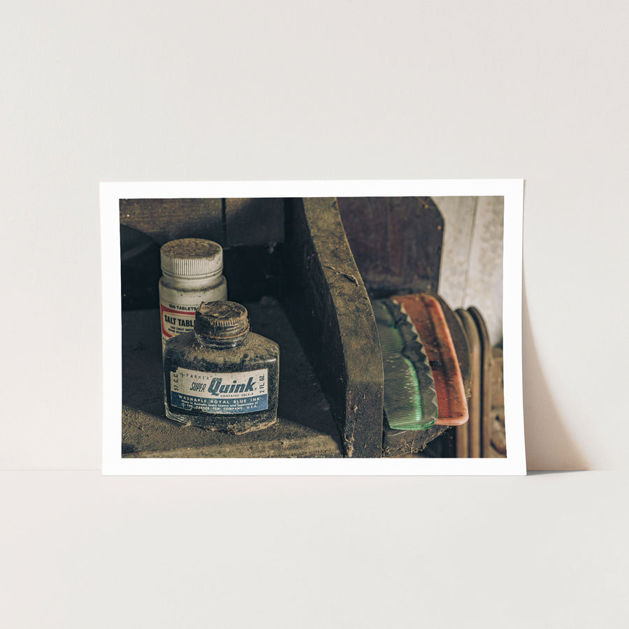 A dust-covered glass ink bottle and white tablet jar on a grimy timber shelf at Woolla, with a green and pink comb resting beside them.