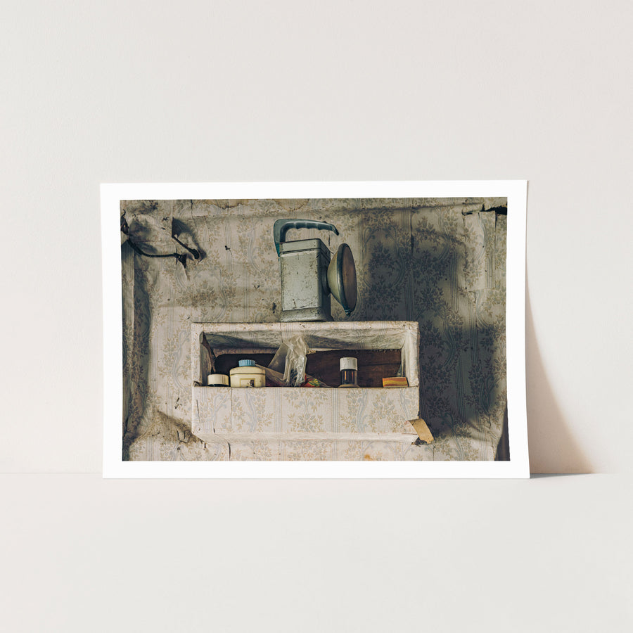 A metal lantern with its reflector open sits on a recessed shelf set into torn floral wallpaper at Woolla, with small jars, a lidded container, a plastic bag, and a matchbox arranged below it.