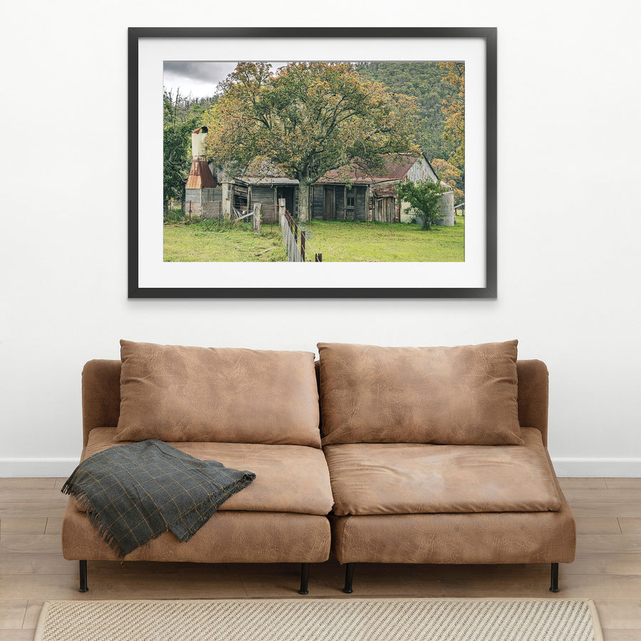 A weathered timber and corrugated iron farmhouse at Woolla, seen behind a large tree in autumn colour, with rusting roof sheets, a tall metal chimney flue, and galvanised tanks, forested hills in the background.