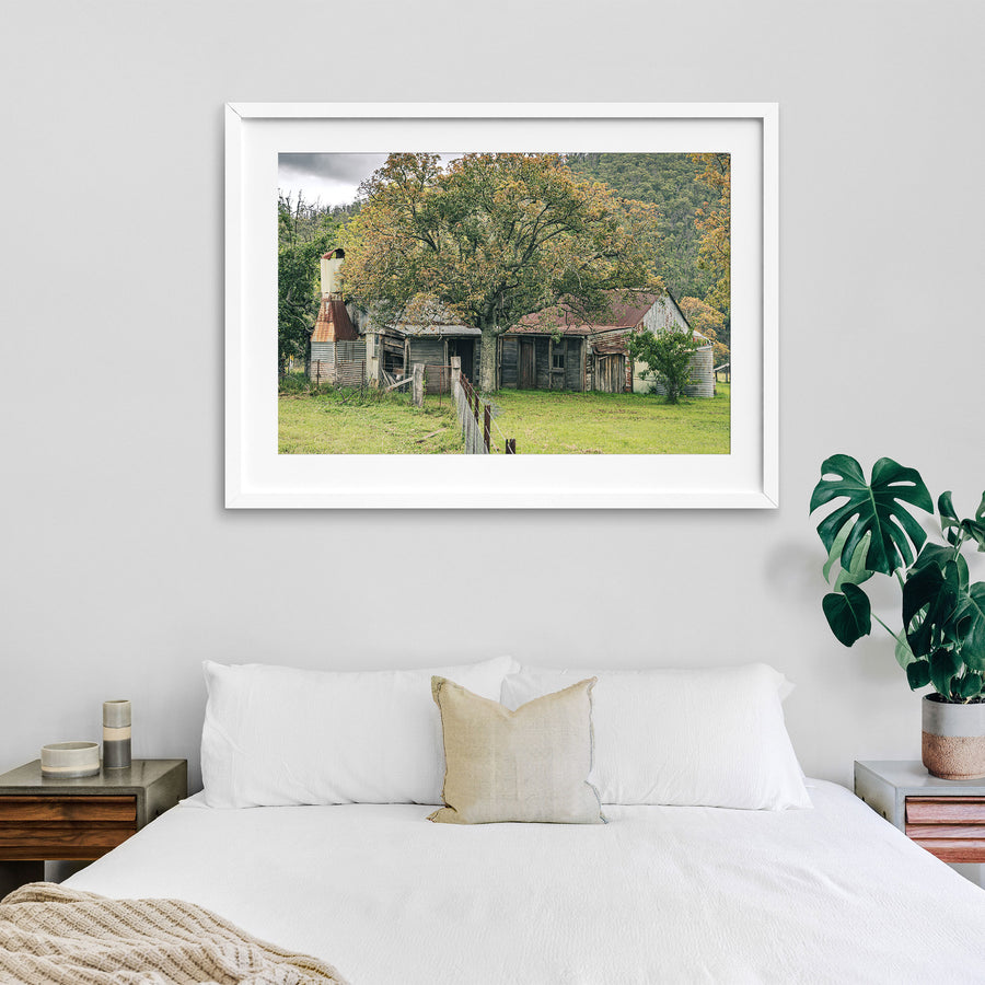 A weathered timber and corrugated iron farmhouse at Woolla, seen behind a large tree in autumn colour, with rusting roof sheets, a tall metal chimney flue, and galvanised tanks, forested hills in the background.
