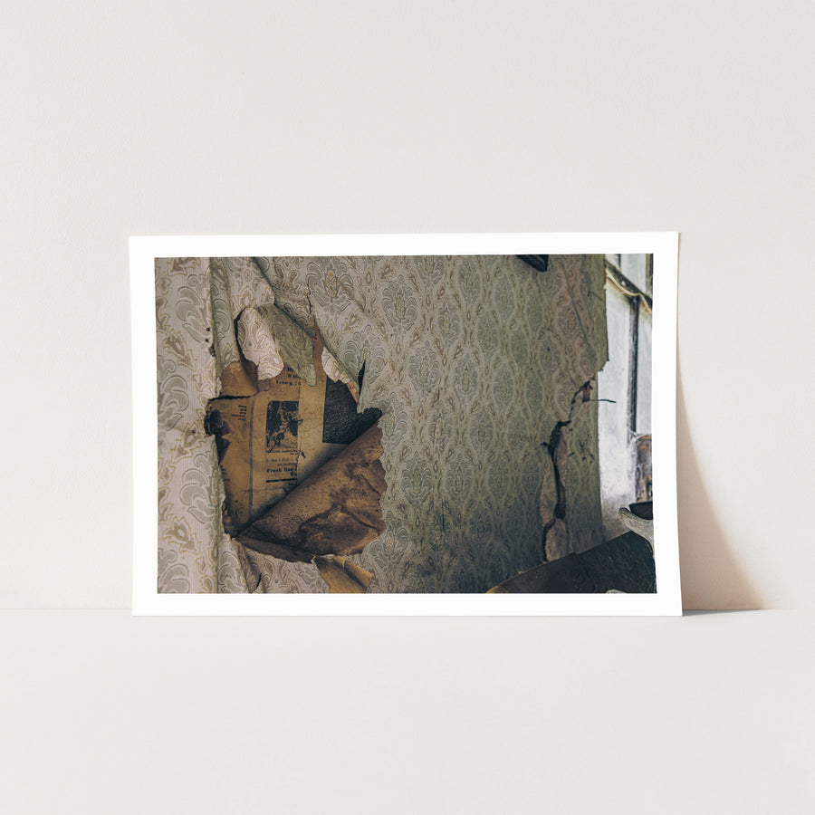 Peeling patterned wallpaper inside a slab hut at Woolla, revealing yellowed newsprint with livestock sale advertising and the partial name Frank Ganz, with a window and damaged plaster visible to the right.