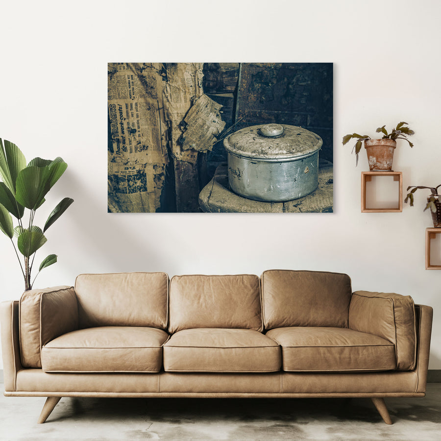 A dust-covered aluminium saucepan and domed lid on a worn wooden surface inside a slab hut at Woolla in the Deua River Valley, with yellowed newspaper lining the timber walls behind.