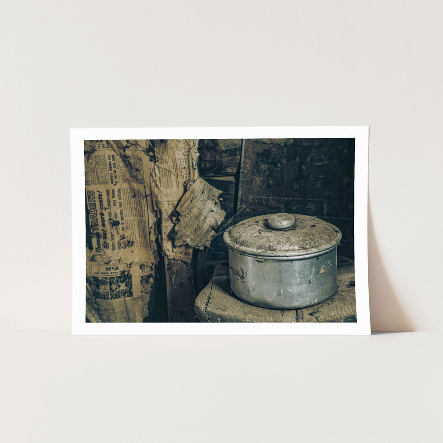 A dust-covered aluminium saucepan and domed lid on a worn wooden surface inside a slab hut at Woolla in the Deua River Valley, with yellowed newspaper lining the timber walls behind.