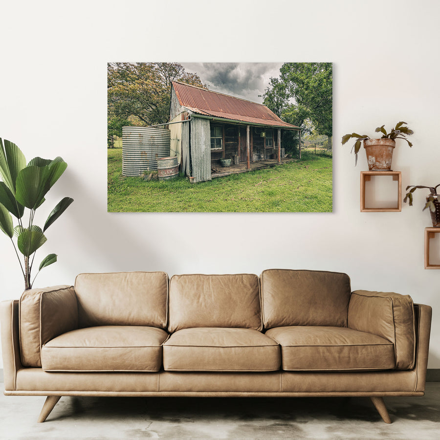 The weathered timber slab hut and covered front verandah at Woolla, with a galvanised corrugated water tank and steel drum standing at the gable end on mown grass among trees near Braidwood.