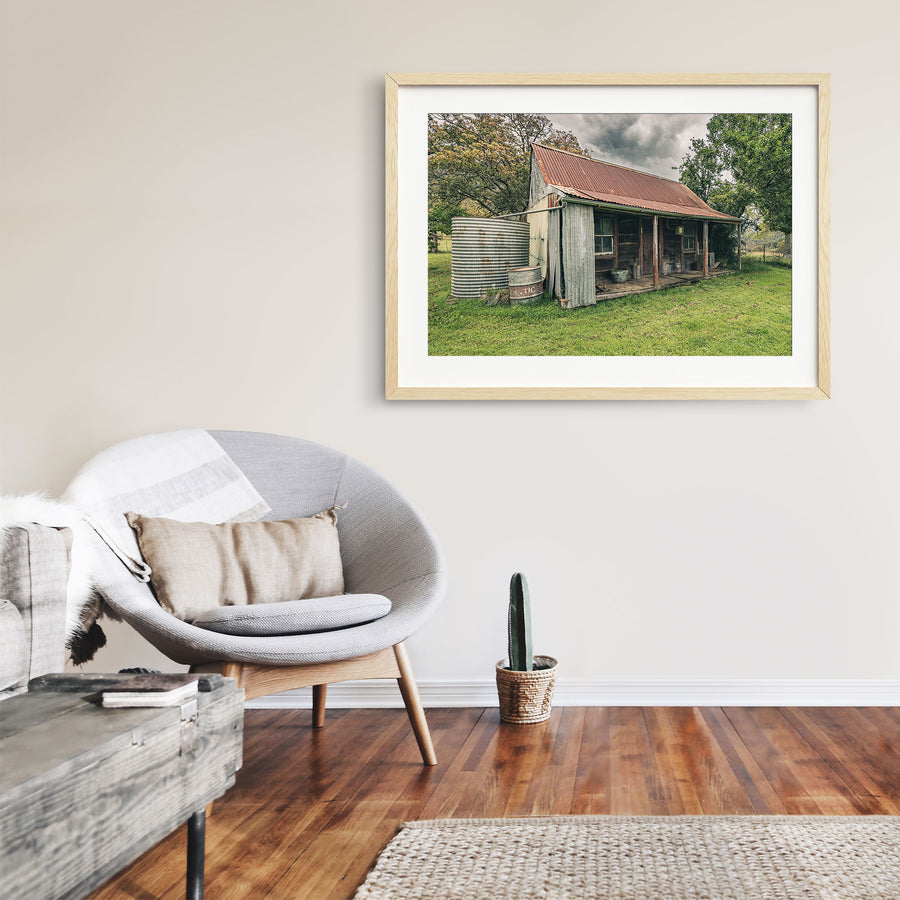The weathered timber slab hut and covered front verandah at Woolla, with a galvanised corrugated water tank and steel drum standing at the gable end on mown grass among trees near Braidwood.
