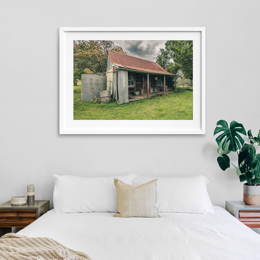 The weathered timber slab hut and covered front verandah at Woolla, with a galvanised corrugated water tank and steel drum standing at the gable end on mown grass among trees near Braidwood.