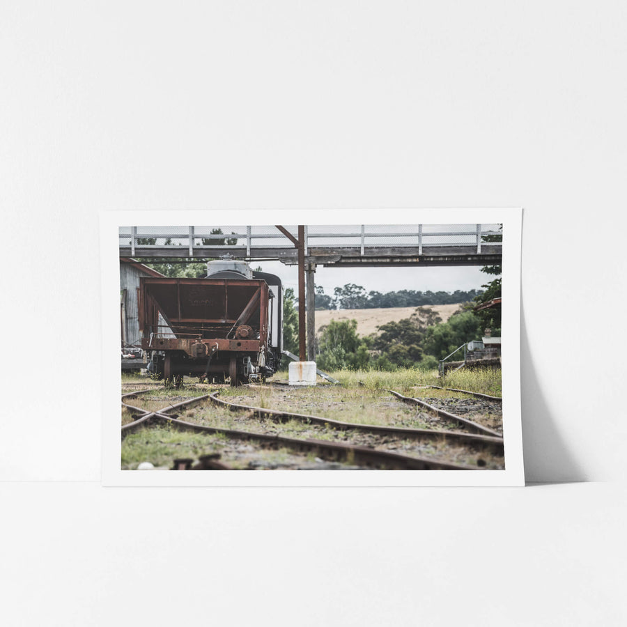Upline at Bombala Station, a rusted hopper wagon sits low on the sidings beneath a steel and timber footbridge.