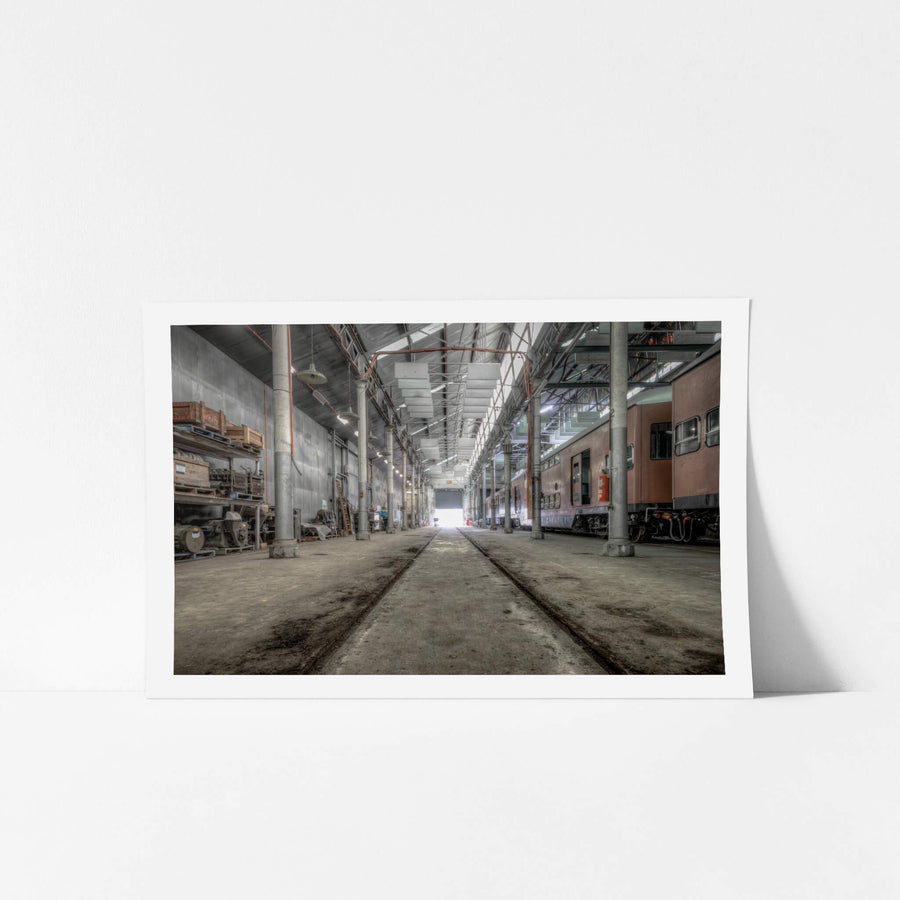 Workshop at Eveleigh Paint Shop, rails run flush with the concrete floor, cutting a straight line through the centre of the workshop toward a bright roller door at the far end.