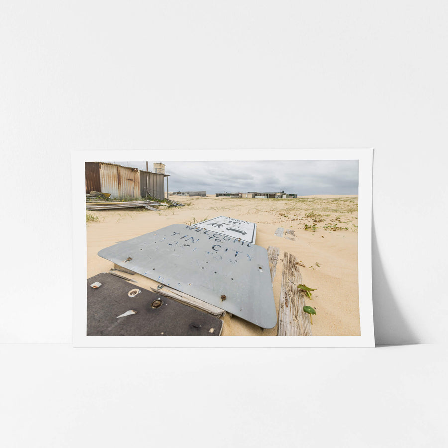 Welcome To Tin City at Tin City, a metal sign lies face-up in the sand, its blue painted letters barely legible.