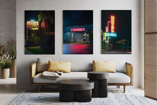 Three Lost Collective neon-sign prints hung above a timber-framed sofa in a styled living room. Left: a motel office sign at night. Centre: a YUMMY diner sign in red neon. Right: a 3 Sisters motel sign.