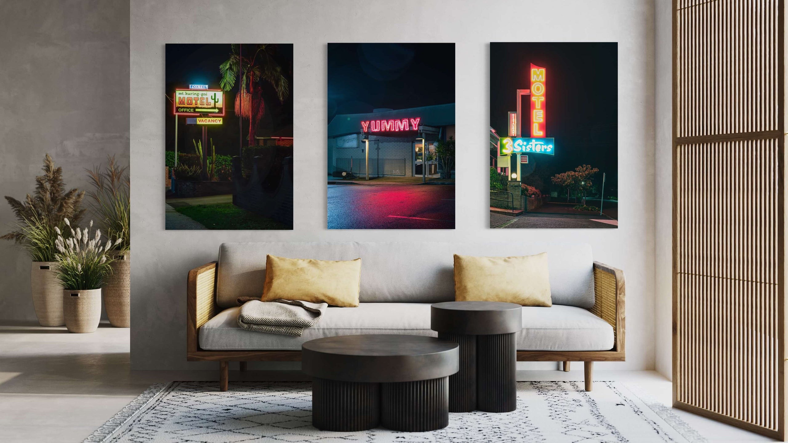 Three Lost Collective neon-sign prints hung above a timber-framed sofa in a styled living room. Left: a motel office sign at night. Centre: a YUMMY diner sign in red neon. Right: a 3 Sisters motel sign.