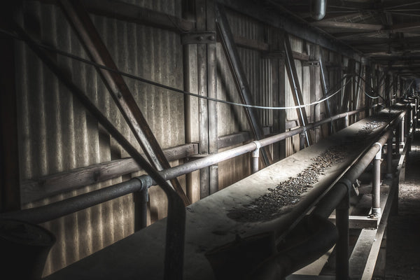 A long pipe-and-conveyor corridor inside the Bradmill mill, lined with corrugated metal walls and diagonal timber bracing, lit by a thin band of light from the far end.
