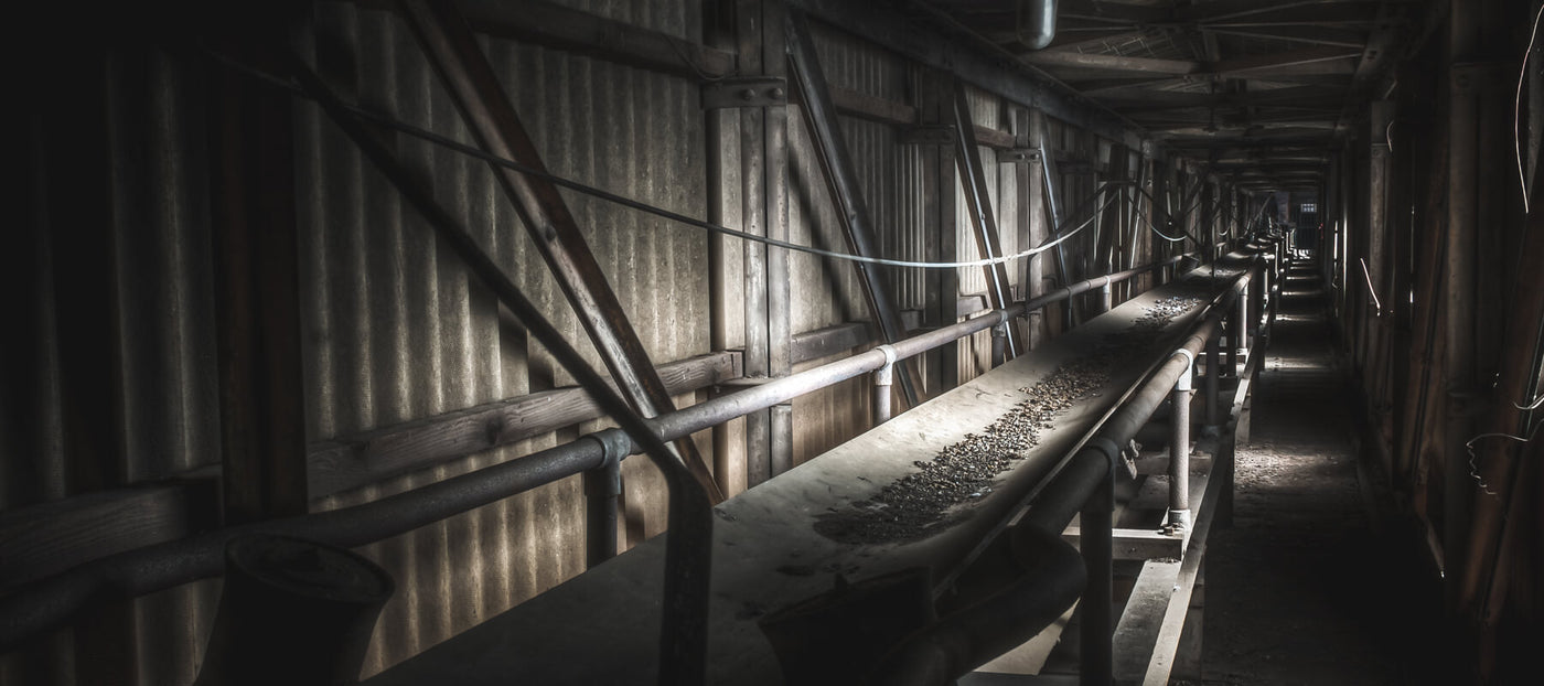 A long pipe-and-conveyor corridor inside the Bradmill mill, lined with corrugated metal walls and diagonal timber bracing, lit by a thin band of light from the far end.