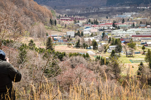 Brett Patman from behind, in a dark hooded jacket, photographing the town of Yubari from a hillside above. The valley below scattered with low buildings and bare winter trees.