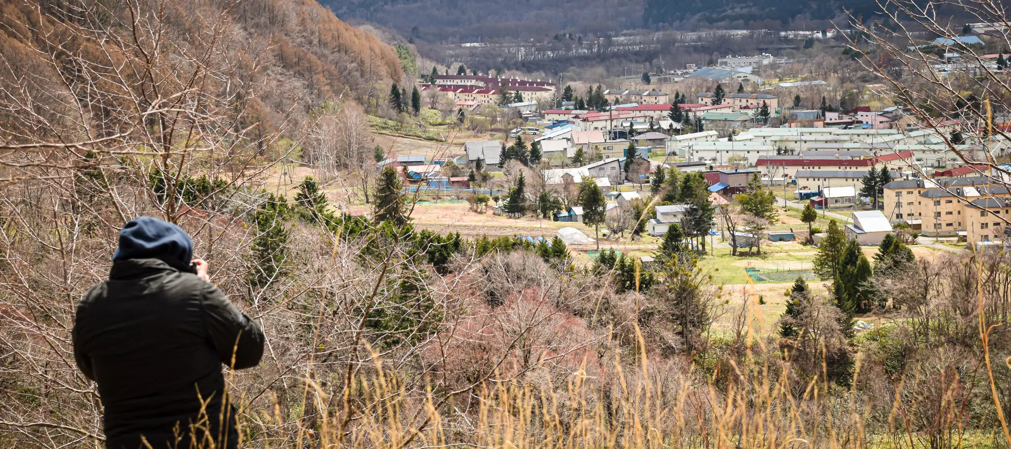 Brett Patman from behind, in a dark hooded jacket, photographing the town of Yubari from a hillside above. The valley below scattered with low buildings and bare winter trees.